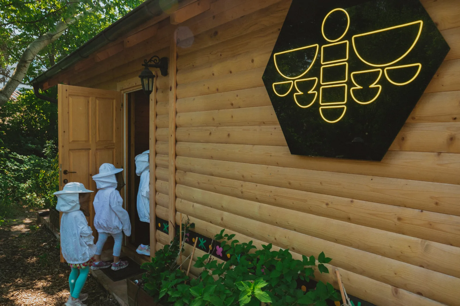 Children in beekeeping suits going into the timber cabin with its neon bee sign