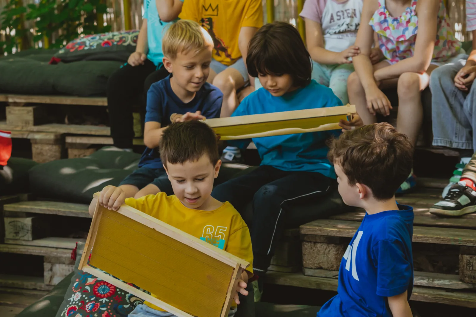 A boy holding a frame of wax foundation at the workshop, the other children sitting around him on pallets