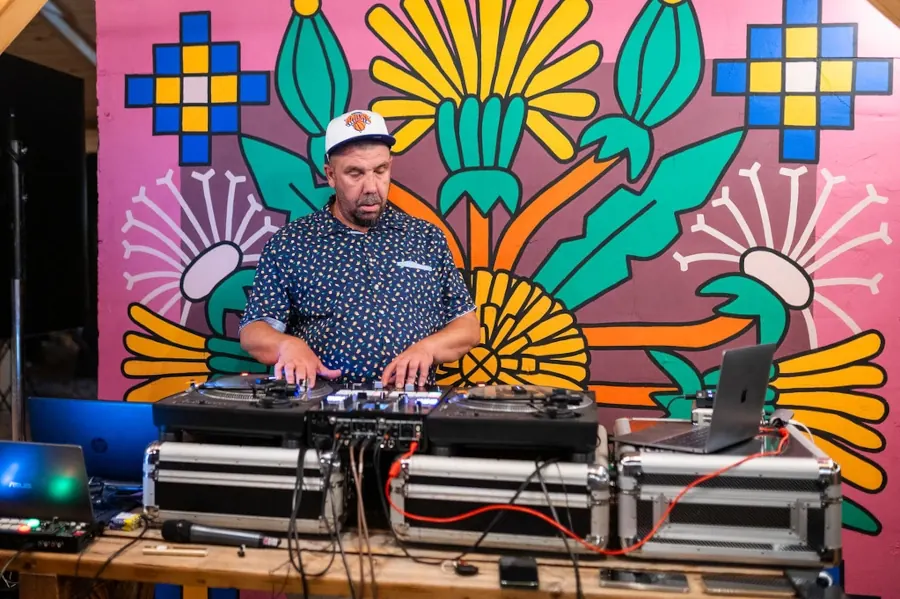 DJ za pultom od paleta ispred ružičastog murala sa cvetnim motivima, uveče u vrtu
