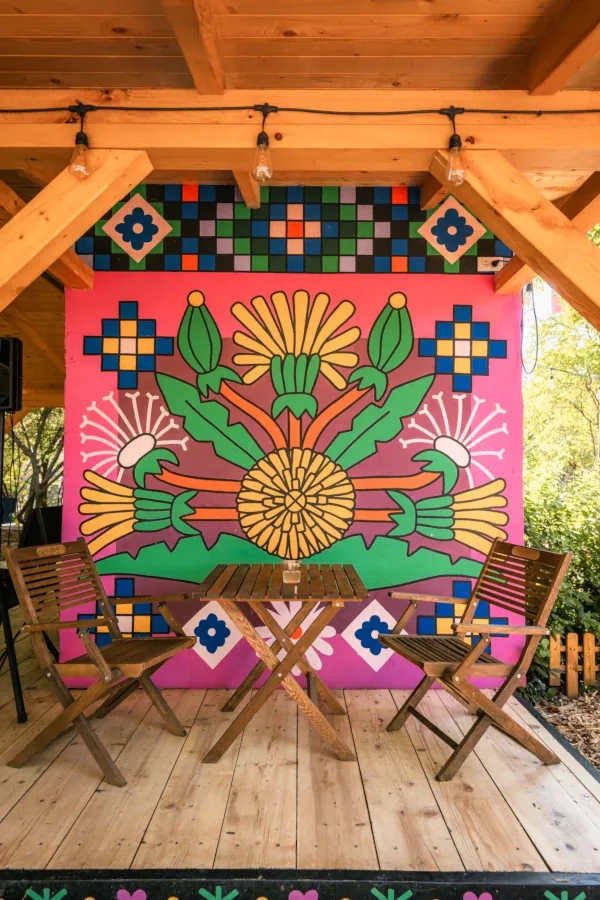 Drveni sto sa dve stolice pod nadstrešnicom, iza njih mural sa cvetnim motivima i sijalice